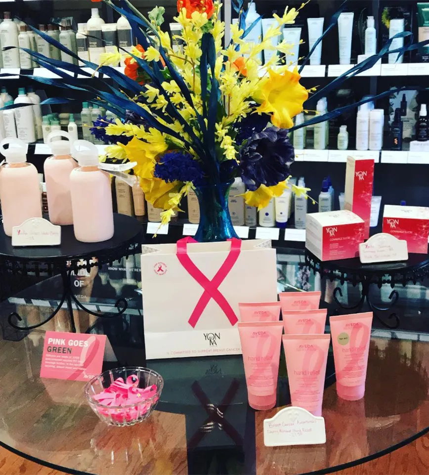 Display of pink skincare products, a gift bag with a pink ribbon, and a bouquet of yellow and blue flowers on a glass table in a store with shelves of beauty products in the background—creating an inviting atmosphere reminiscent of a luxurious day spa Covington GA or an upscale hair salon and spa Newton County. - La Vie Spa & Salon in Covington, GA