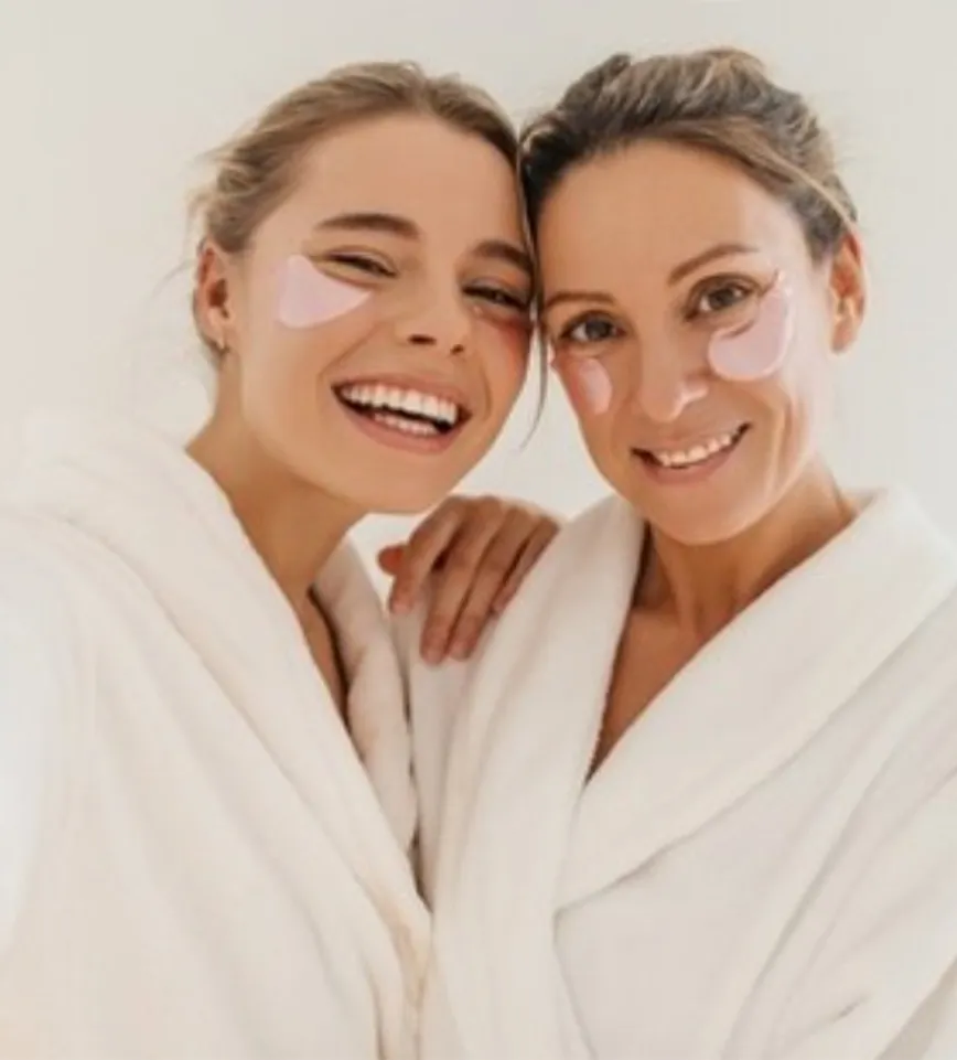Two women wearing white robes and pink under-eye patches smile at the camera, standing close together against a plain background—capturing a moment of relaxation after Yon-Ka Paris facial treatments at La Vie Spa & Salon. - La Vie Spa & Salon in Covington, GA