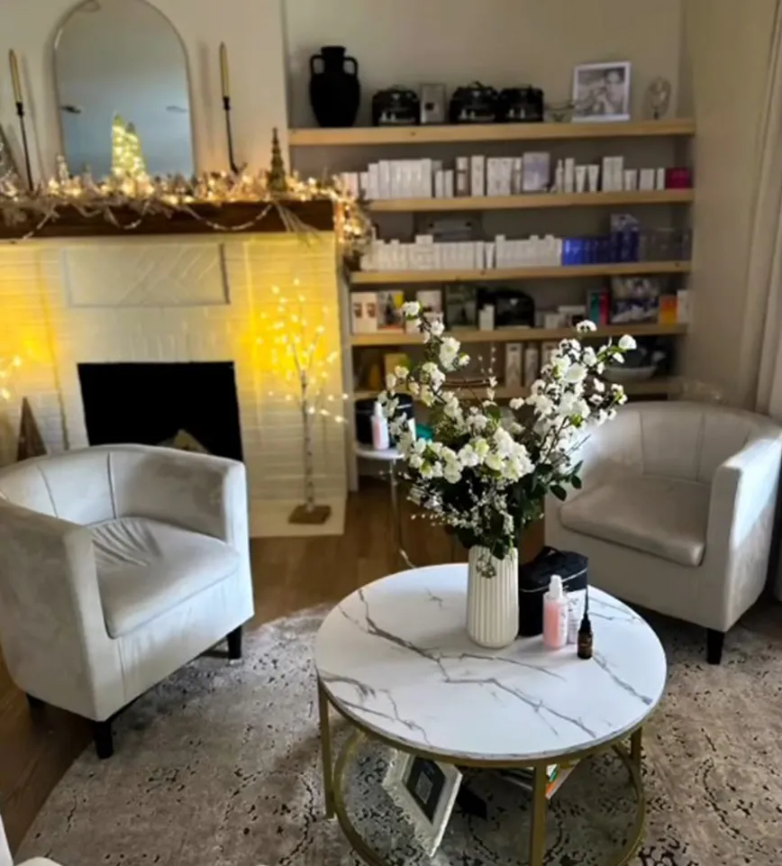 A well-lit room with two armchairs, a round marble coffee table, a decorated mantelpiece with string lights, and shelves holding bottles and boxes evokes the tranquil atmosphere of a hair salon and spa in Newton County, inviting guests to unwind in style. - La Vie Spa & Salon in Covington, GA