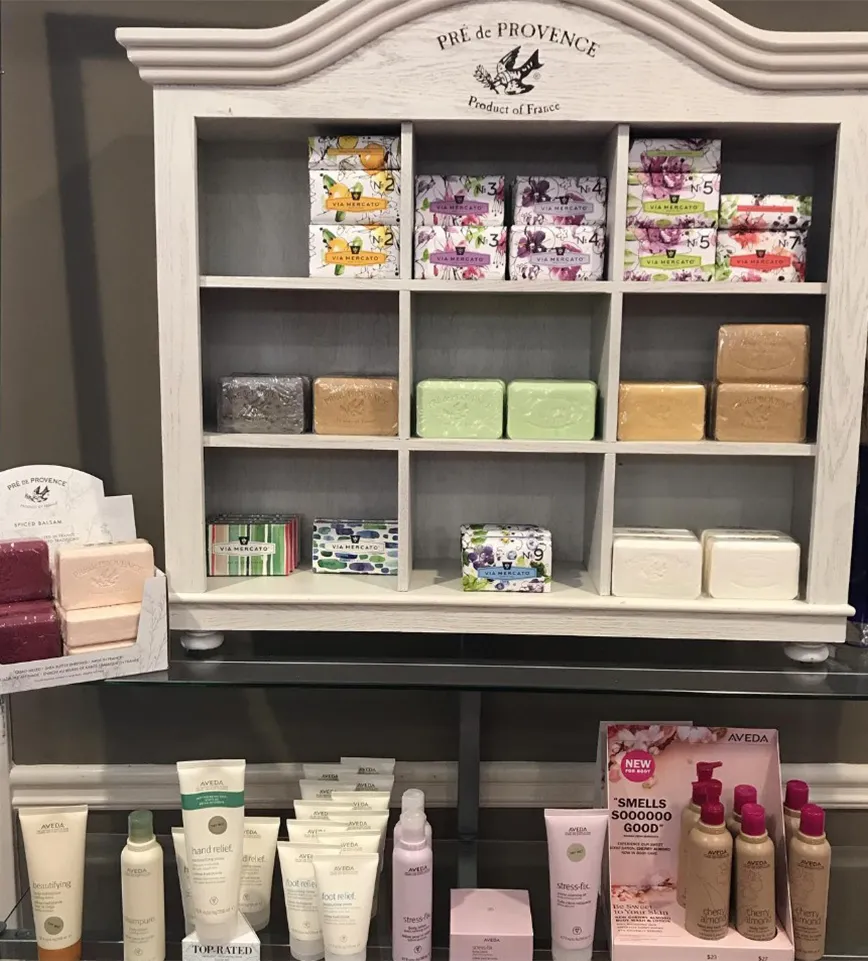 White shelf displays assorted boxed and unboxed soaps, with several skincare products and lotions arranged on the counter below. Signage indicates brands Pré de Provence and Aveda, reflecting the luxurious offerings you’ll find at La Vie Spa & Salon, a premier day spa in Covington GA known for Yon-Ka Paris facial treatments. - La Vie Spa & Salon in Covington, GA