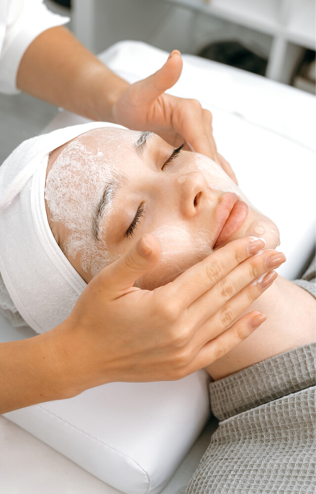 Person lying with eyes closed, receiving a facial treatment as foam cleanser is applied to the face by another person’s hands in a soothing day spa setting at La Vie Spa & Salon, Covington GA—experience the luxurious Yon-Ka Paris facial treatments designed for ultimate relaxation and radiant skin. - La Vie Spa & Salon in Covington, GA