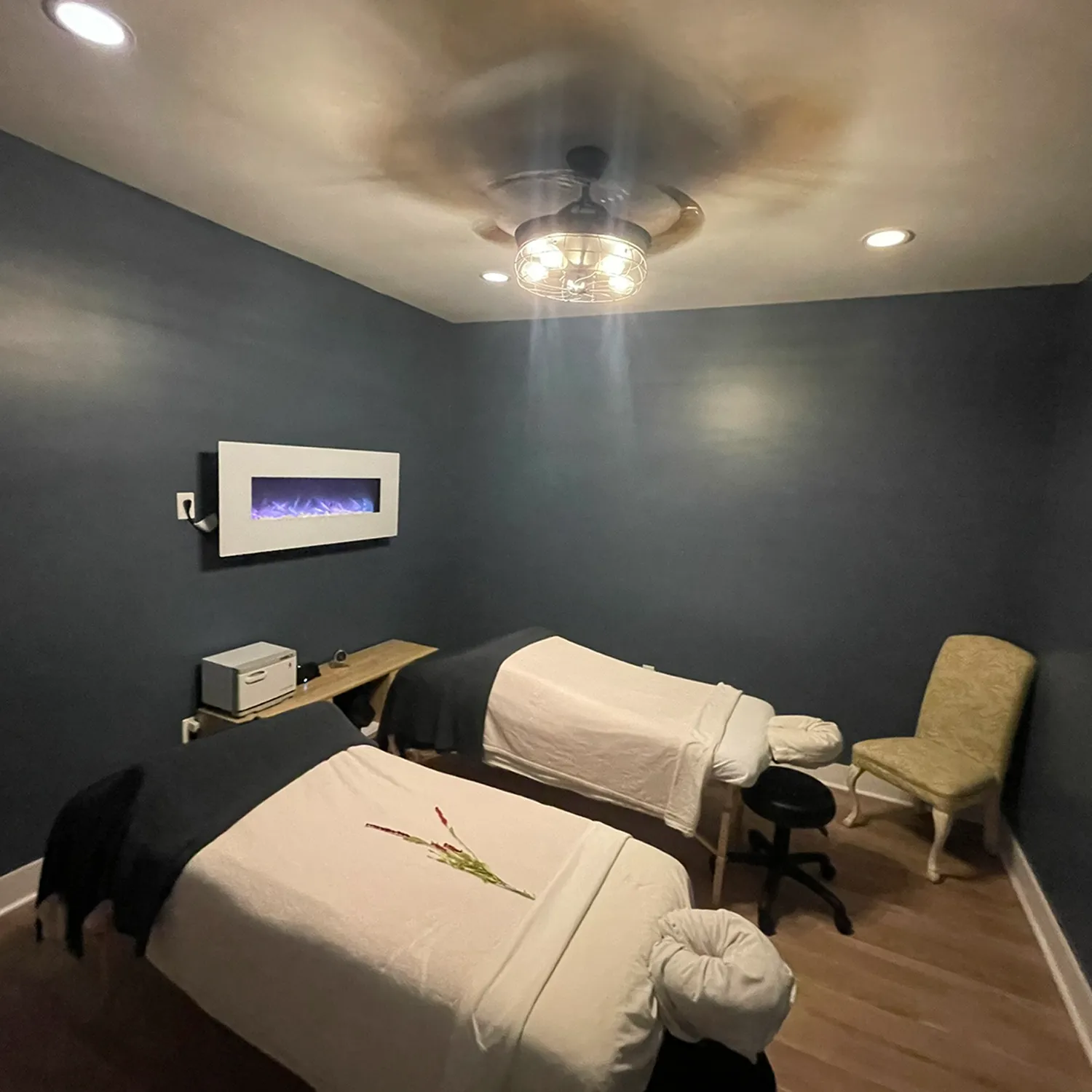 Two massage tables with white linens and a flower decoration await in a dimly lit room with dark walls, featuring a wall-mounted electric fireplace, a chair, and a small shelf—the perfect setting to experience deep tissue massage therapy or indulge in Yon-Ka Paris facial treatments at our hair salon and spa in Newton County. - La Vie Spa & Salon in Covington, GA