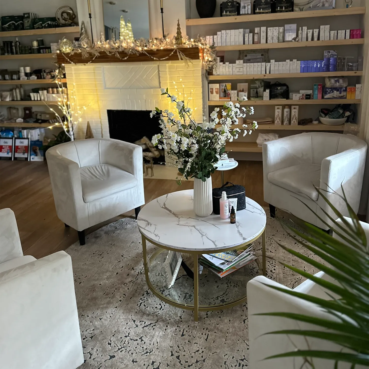 A cozy room with four white armchairs around a marble coffee table, a vase of flowers, shelves with products, and a decorated fireplace in the background—perfect for relaxing before your Yon-Ka Paris facial treatments at this inviting day spa in Covington, GA. - La Vie Spa & Salon in Covington, GA
