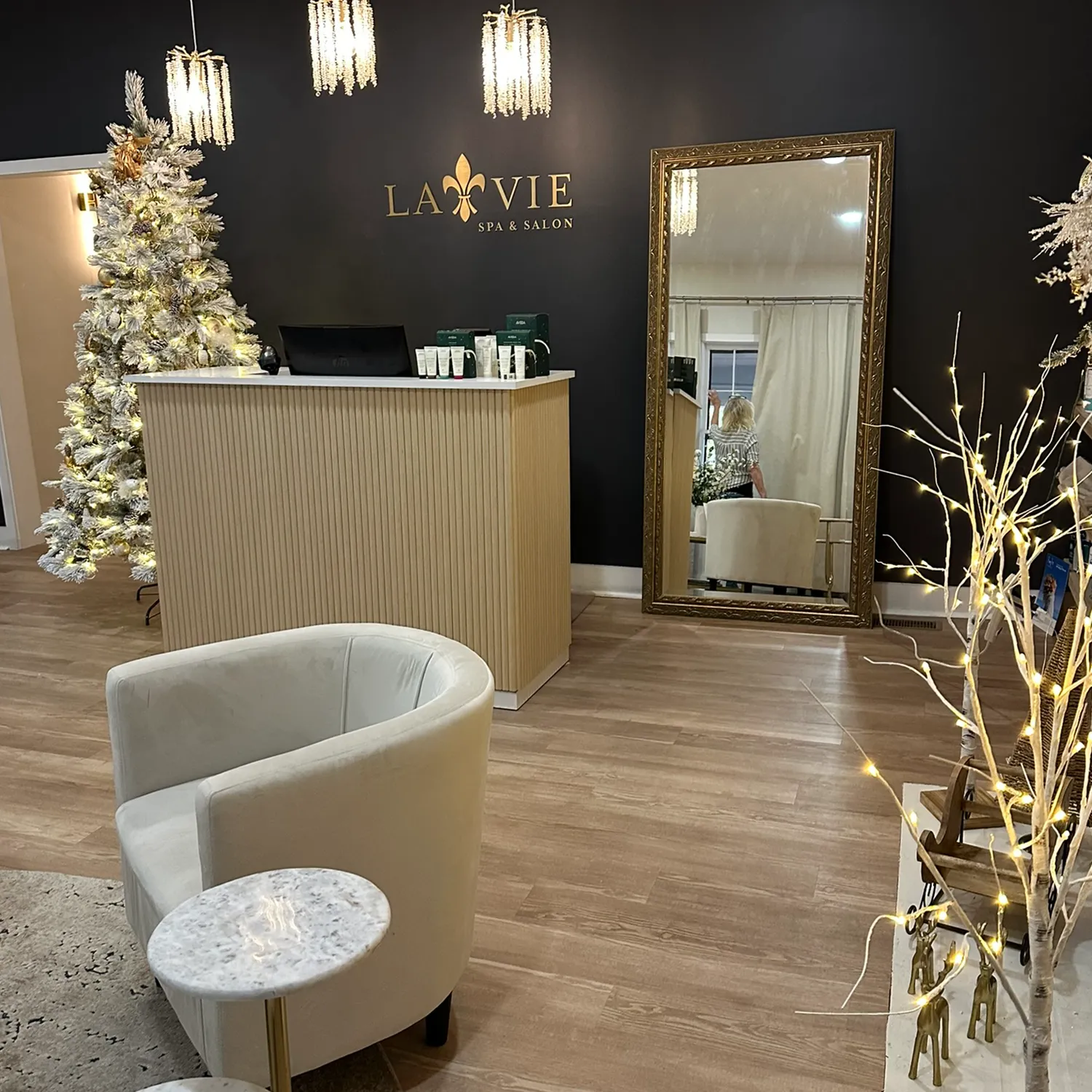 Reception area of La Vie Spa & Salon, a welcoming day spa in Covington, GA, featuring a beautifully decorated Christmas tree, large mirror, white chair, small round table, and sparkling holiday lights against a dark accent wall with “La Vie Spa & Salon” signage—an inviting space perfect for relaxing before your deep tissue massage therapy. - La Vie Spa & Salon in Covington, GA