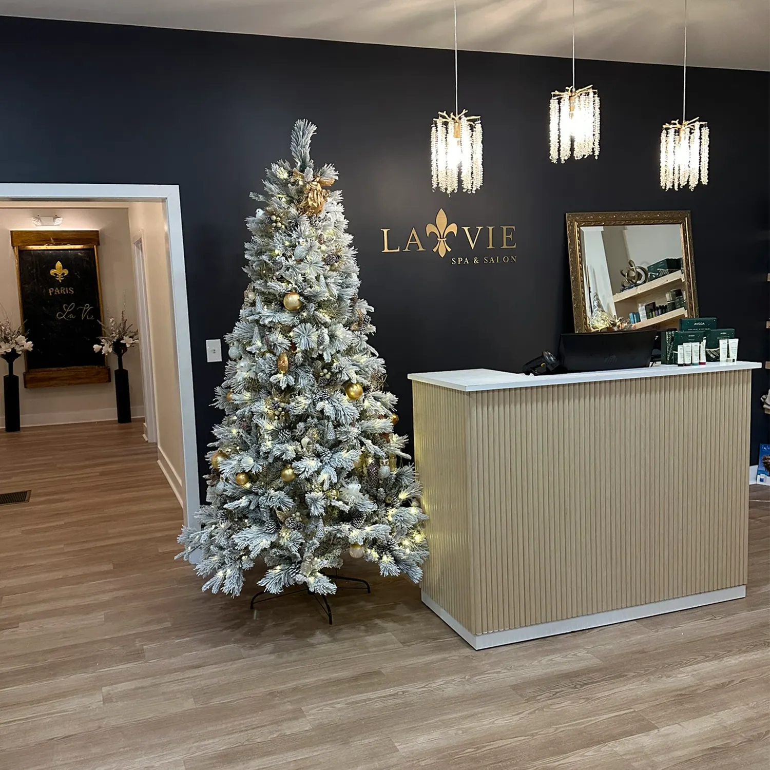 Reception area of La Vie Spa & Salon, a premier hair salon and spa in Newton County, featuring a stunning white Christmas tree adorned with gold ornaments, modern pendant lights, a wooden floor, and a welcoming front desk showcasing Yon-Ka Paris facial treatments alongside other premium beauty products. - La Vie Spa & Salon in Covington, GA