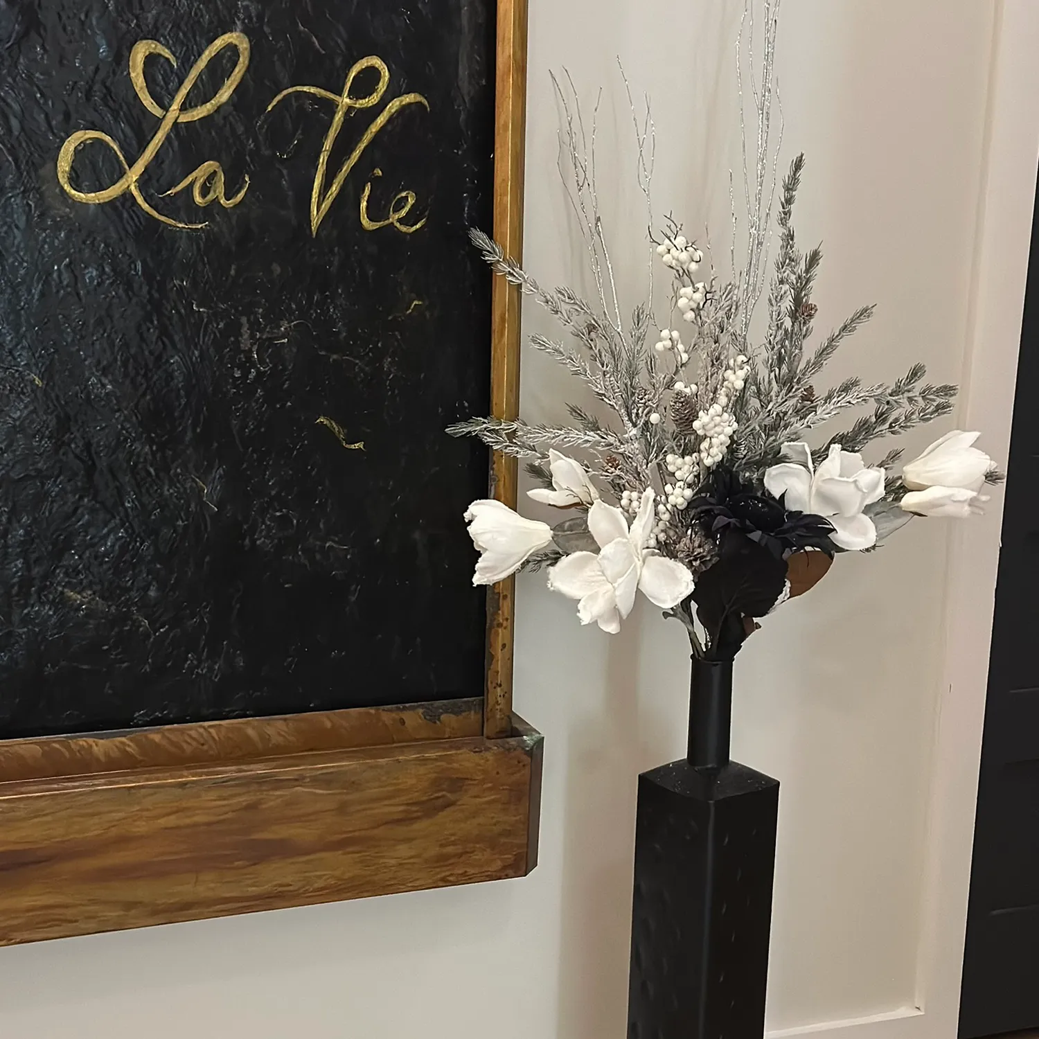 A black vase with white and gray artificial flowers stands beside a blackboard with “La Vie” written in gold script on a textured black background with a wooden frame, evoking the tranquil atmosphere of a hair salon and spa in Newton County. - La Vie Spa & Salon in Covington, GA