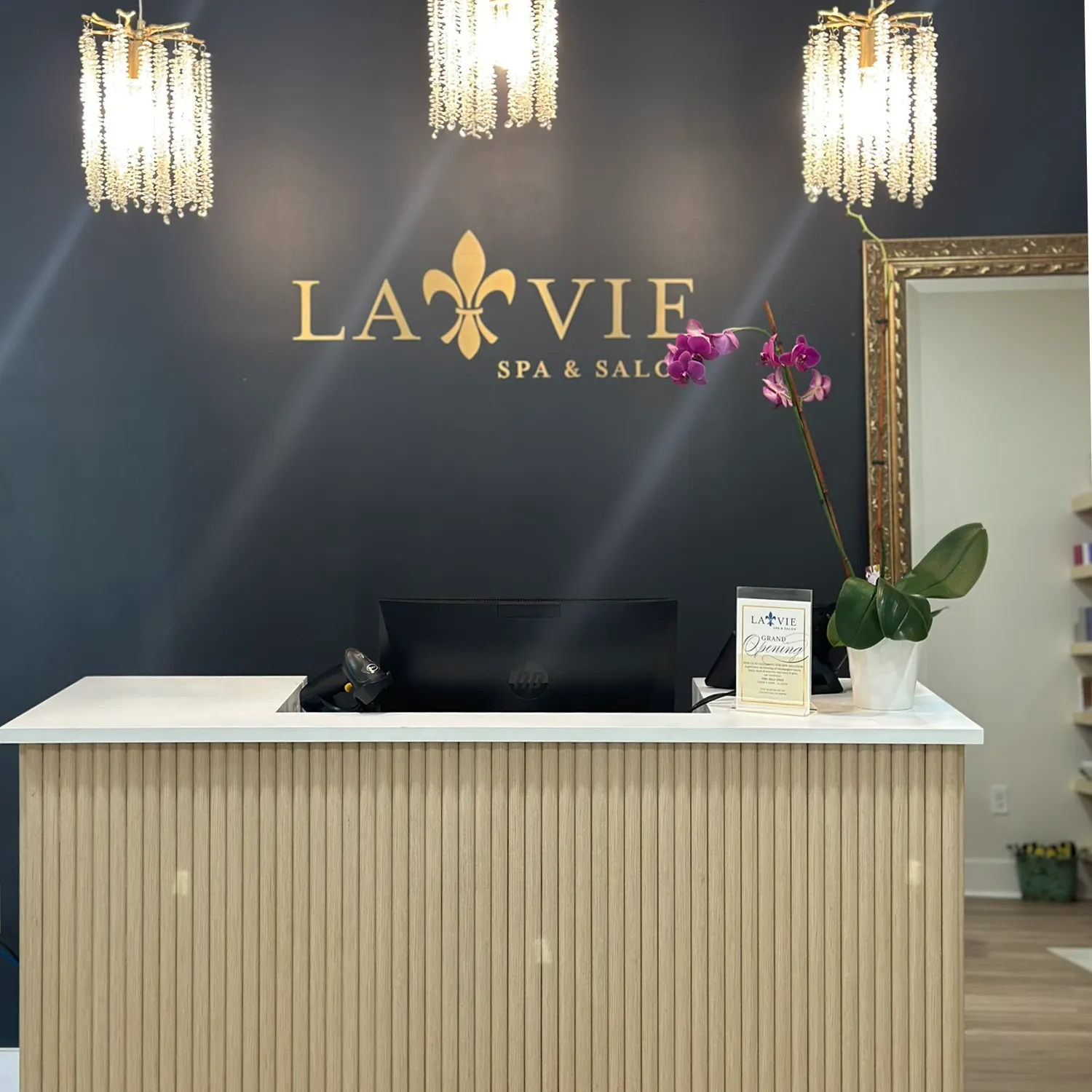 Reception desk at La Vie Spa & Salon—a premier hair salon and spa in Newton County—with a computer, an orchid, a framed sign, and gold pendant lights against a dark wall featuring the logo. - La Vie Spa & Salon in Covington, GA