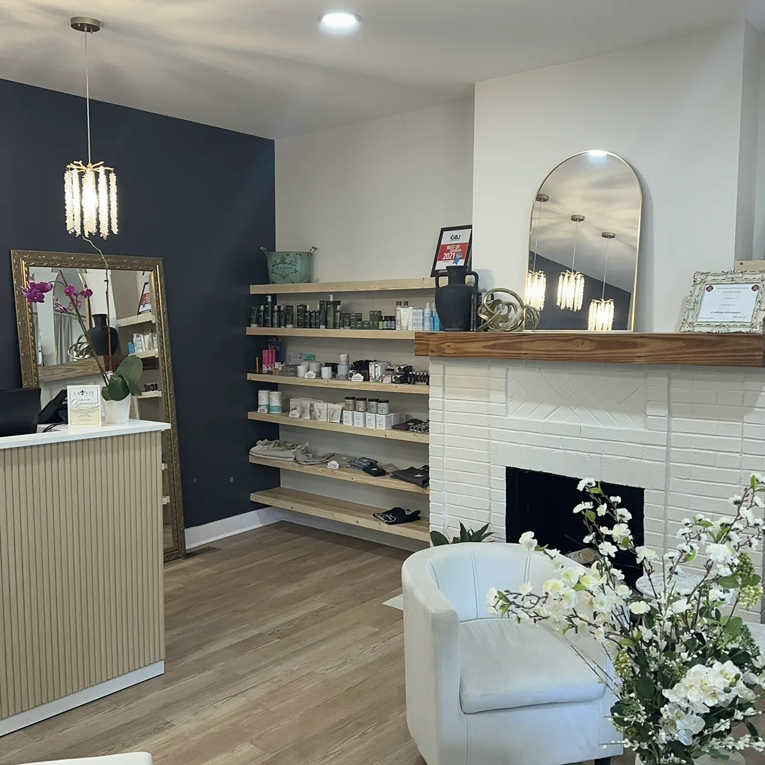 A modern salon reception area with a white counter, shelves of beauty products—including Yon-Ka Paris facial treatments—a fireplace, a round mirror, and decorative flowers welcomes guests to this premier hair salon and spa in Newton County. - La Vie Spa & Salon in Covington, GA
