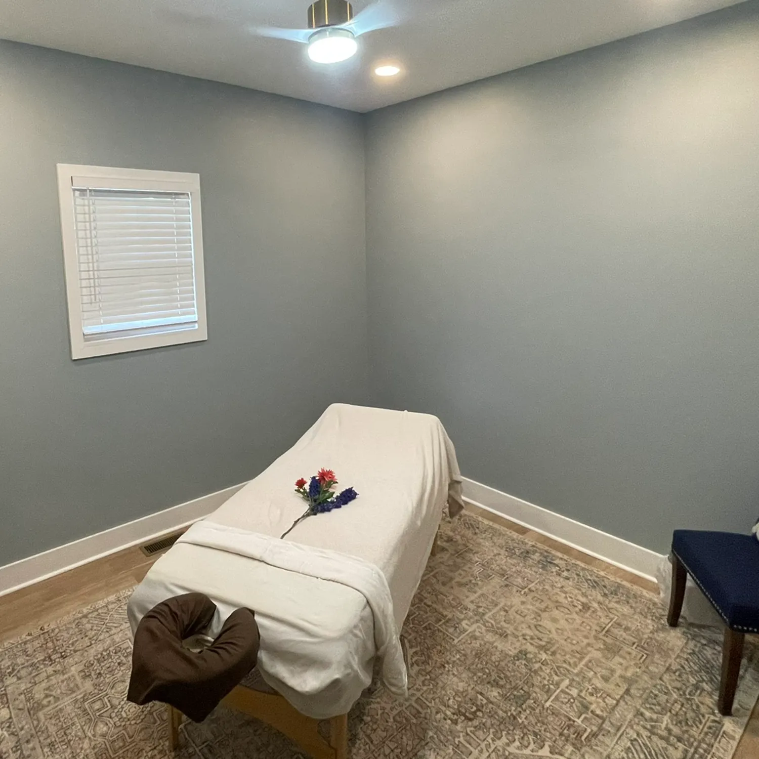 A massage table with white sheets and a small bouquet is set up in a simple room with gray walls, a window, and a chair, creating the calming atmosphere you can expect at La Vie Spa & Salon—your premier day spa in Covington, GA. - La Vie Spa & Salon in Covington, GA